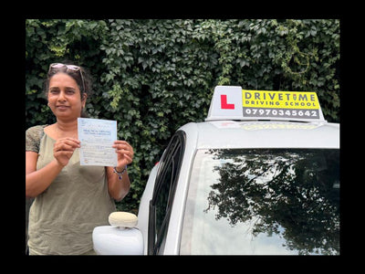 A woman stands outdoors between two cars, holding a document. The car next to her has a yellow 'L' learner license sign on top, and another car is partially visible on the left.
