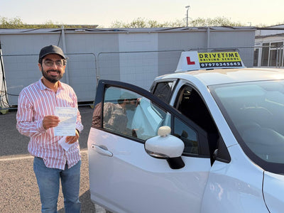 A man in a striped shirt and jeans stands next to a white driving test car with the door open, holding a paper while outdoors near a building and portable toilets.
