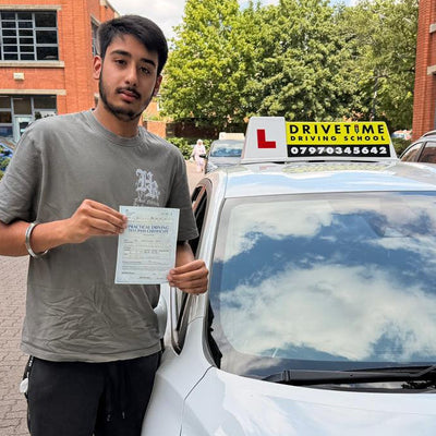 A man in a grey t-shirt and black pants stands next to a white car in an outdoor parking lot, holding a document. The car has an 'L' license plate sign on top. he passed with drivetime first time 3 minors